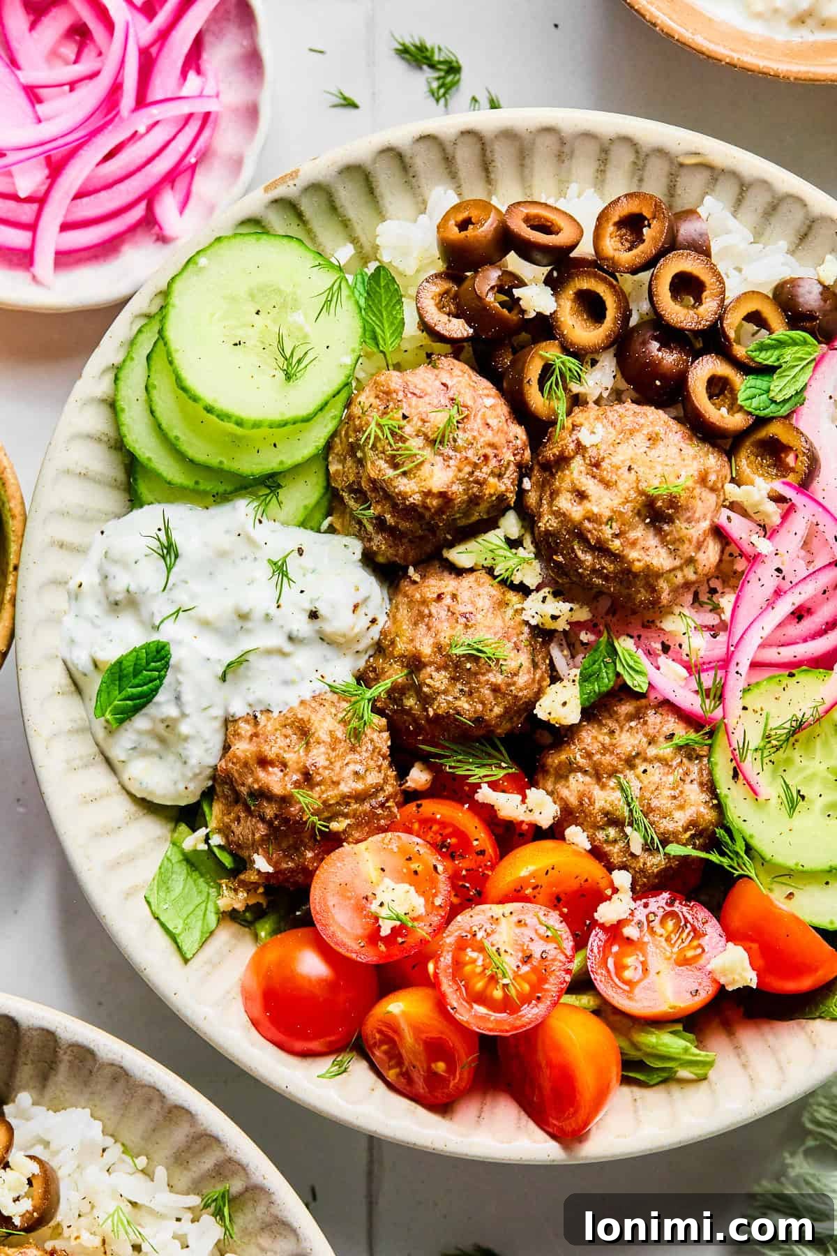 Easy Greek Lamb Meatballs with Zesty Lemon Dill Yogurt 2 Close-up of Greek lamb meatballs served over rice, surrounded by sliced cucumber, black olives, red onion, cherry tomatoes, fresh herbs, and crumbled feta cheese, with lemon dill yogurt sauce on the side.