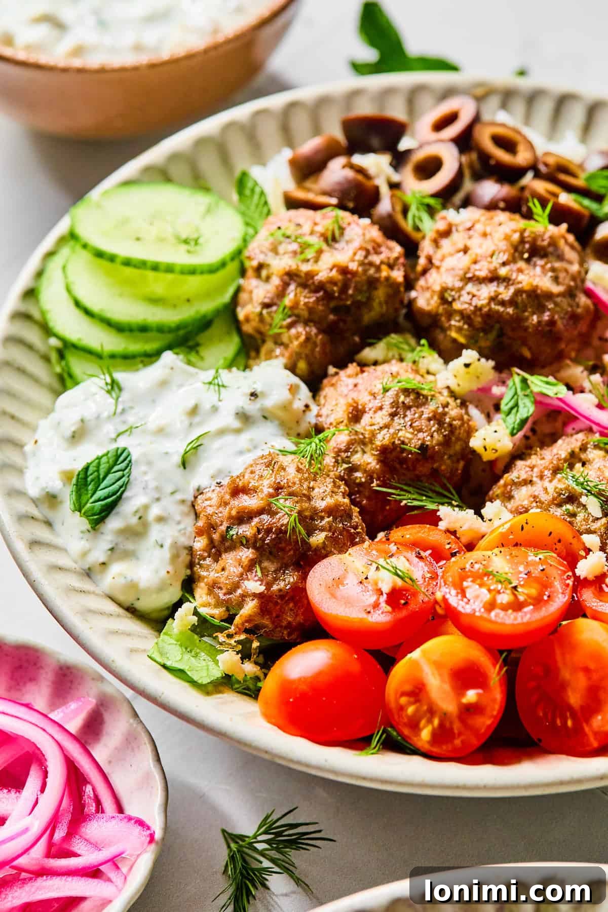 Easy Greek Lamb Meatballs with Zesty Lemon Dill Yogurt 11 Side view of Greek lamb meatballs served over rice, surrounded by sliced cucumber, black olives, red onion, cherry tomatoes, fresh herbs, and crumbled feta cheese, with lemon dill yogurt sauce.