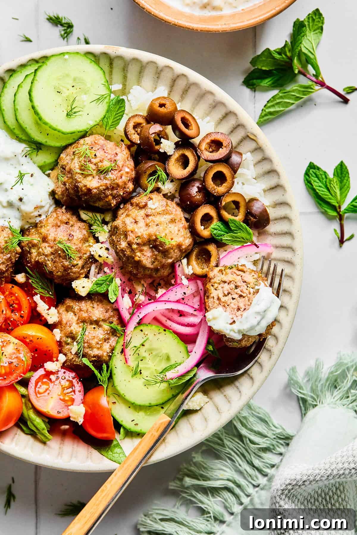 Easy Greek Lamb Meatballs with Zesty Lemon Dill Yogurt 9 Greek lamb meatballs served over rice, surrounded by sliced cucumber, black olives, red onion, cherry tomatoes, fresh herbs, and crumbled feta cheese, with lemon dill yogurt sauce.
