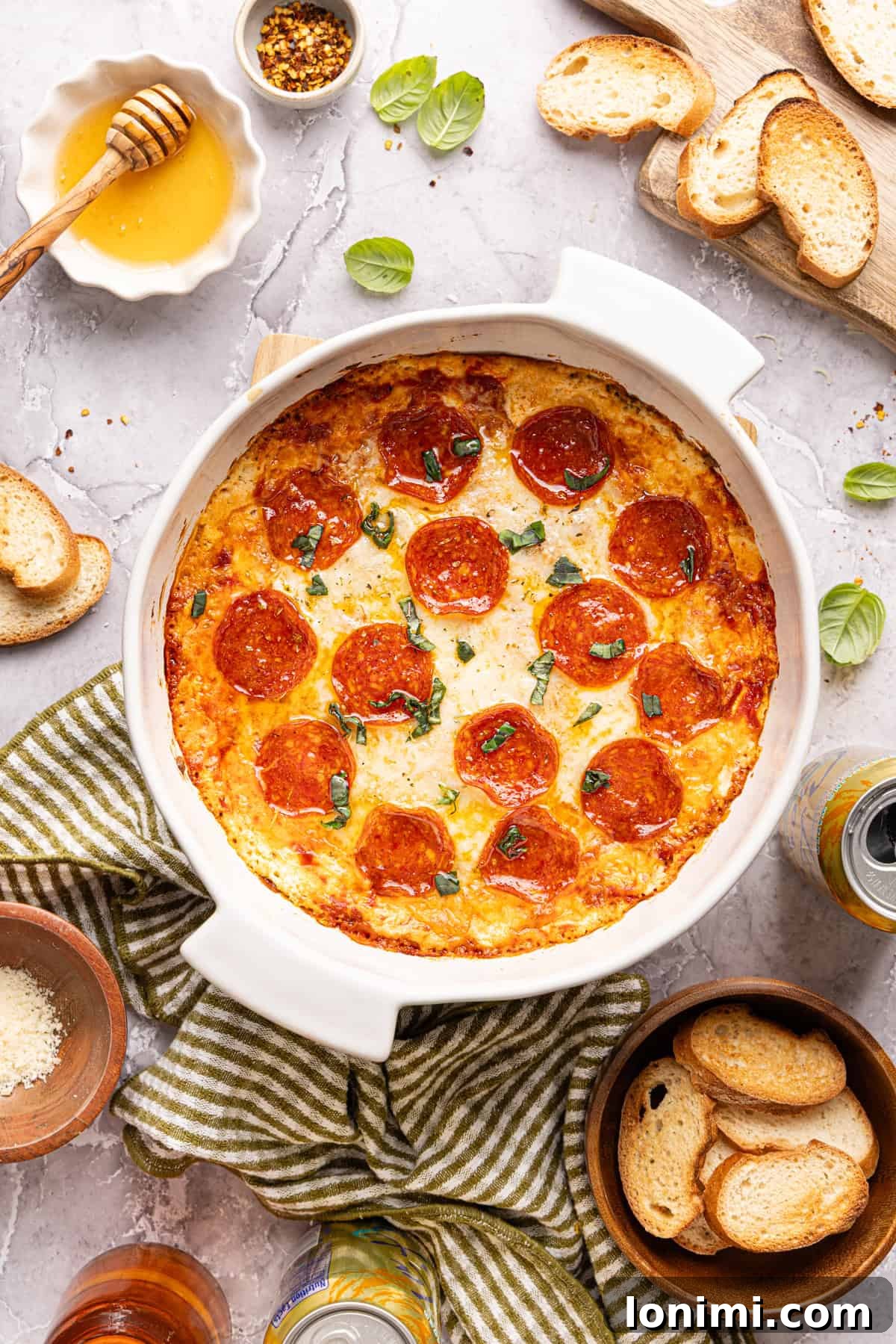 The fully baked high protein pizza dip showcased in a round white dish, garnished with pepperoni, melted cheese, a hot honey drizzle, and fresh basil leaves. Toasted baguette slices are elegantly placed within the dip, ready for serving.
