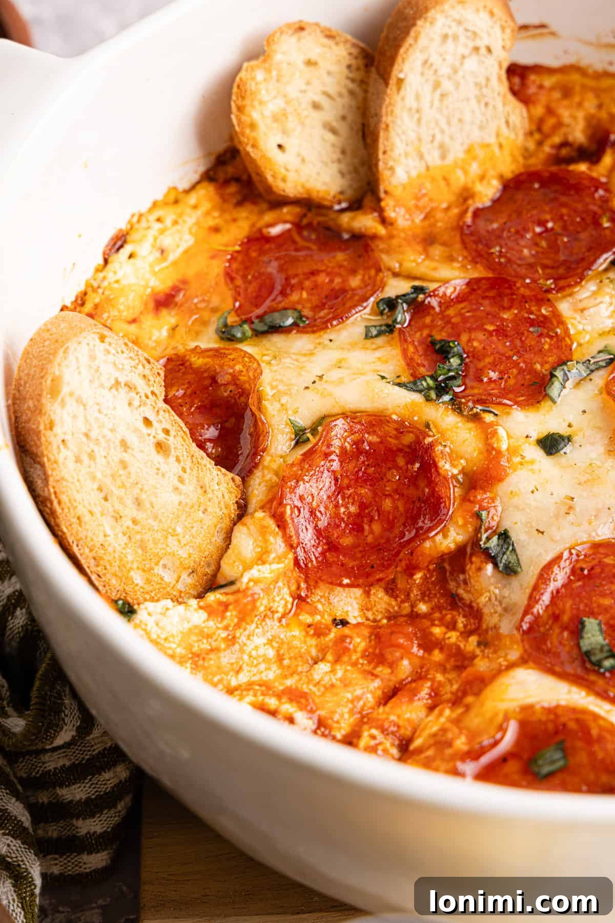 Close-up, appetizing view of the baked high protein pizza dip in a white round dish, generously topped with crispy pepperoni, rich melted cheese, a glistening drizzle of hot honey, and vibrant chopped fresh basil. Several small, golden slices of bread are nestled invitingly within the dip, ready for indulgence.