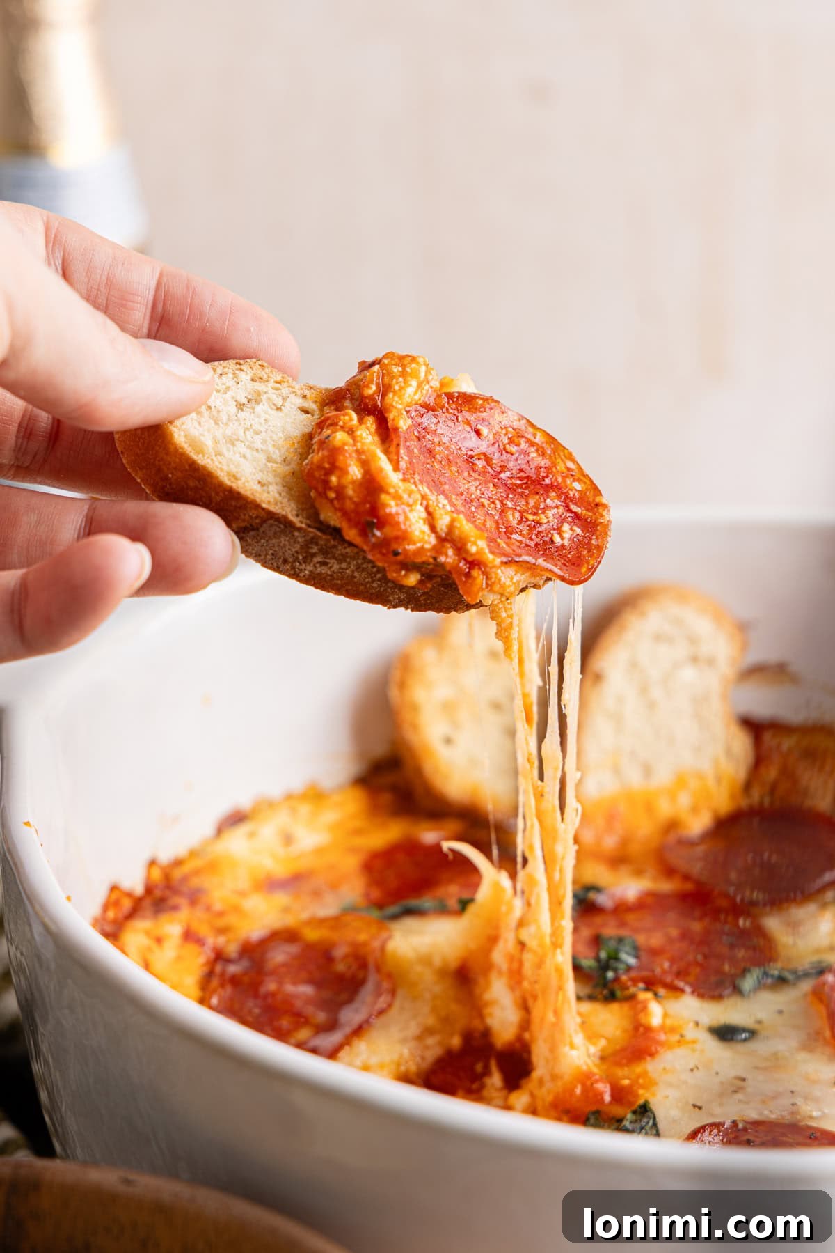A hand holding a perfectly scooped slice of toasted bread, laden with warm, bubbly high protein pizza dip, positioned above the main baking dish filled with the rest of the delicious dip.