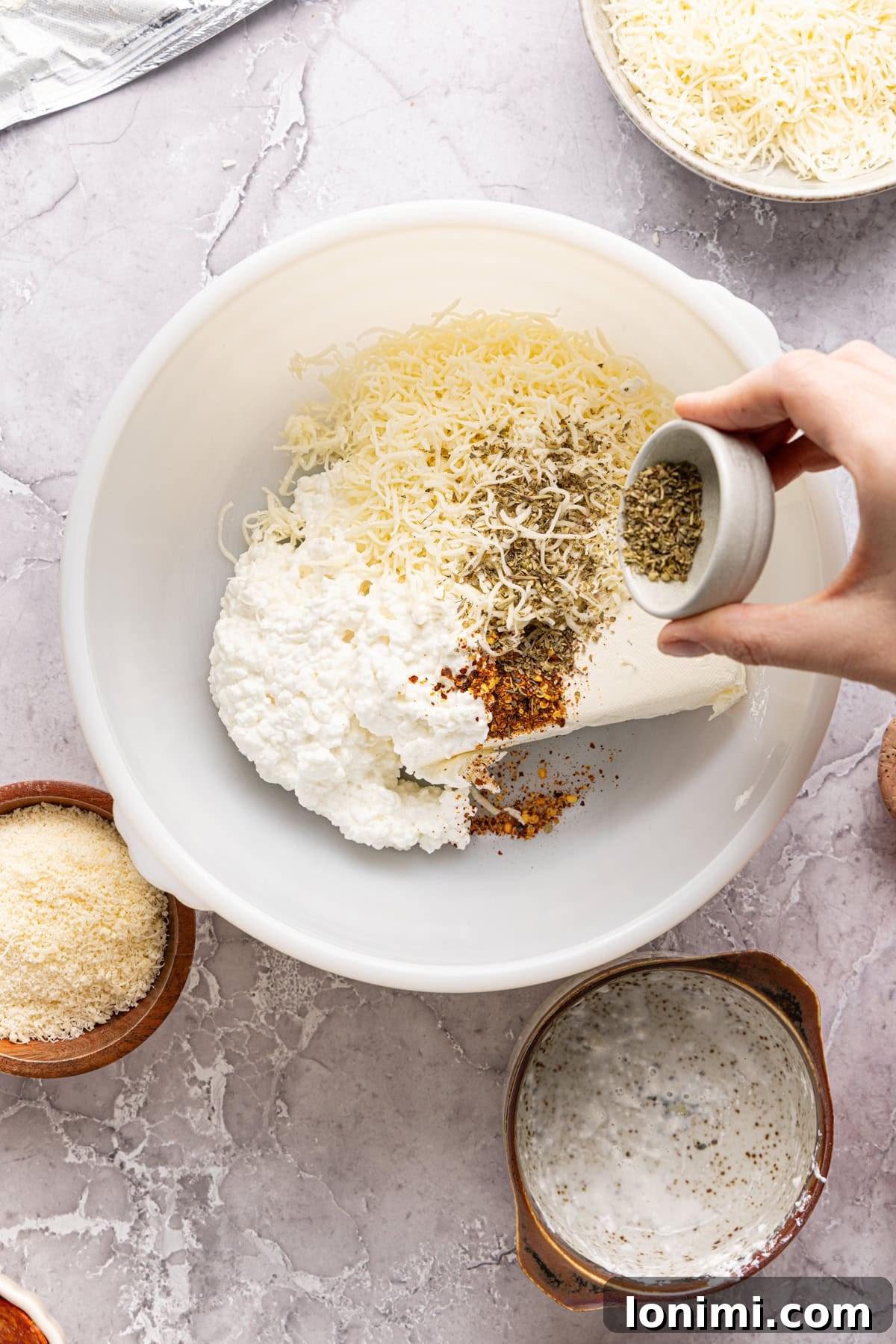White mixing bowl containing the blended base mixture of cream cheese, cottage cheese, mozzarella, parmesan, and seasonings, with a handheld electric mixer resting inside, ready for action.