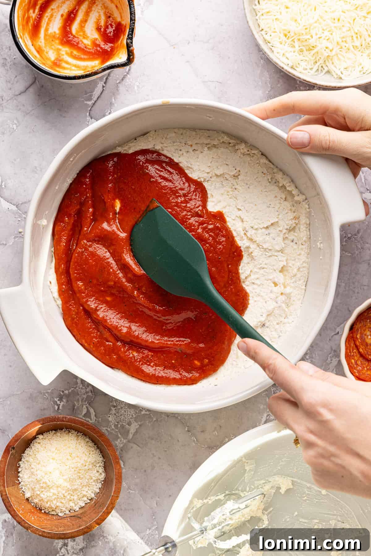 A spoon carefully spreading a vibrant red layer of pizza sauce over the evenly distributed white cheese mixture in a baking dish, creating the next layer of the dip.