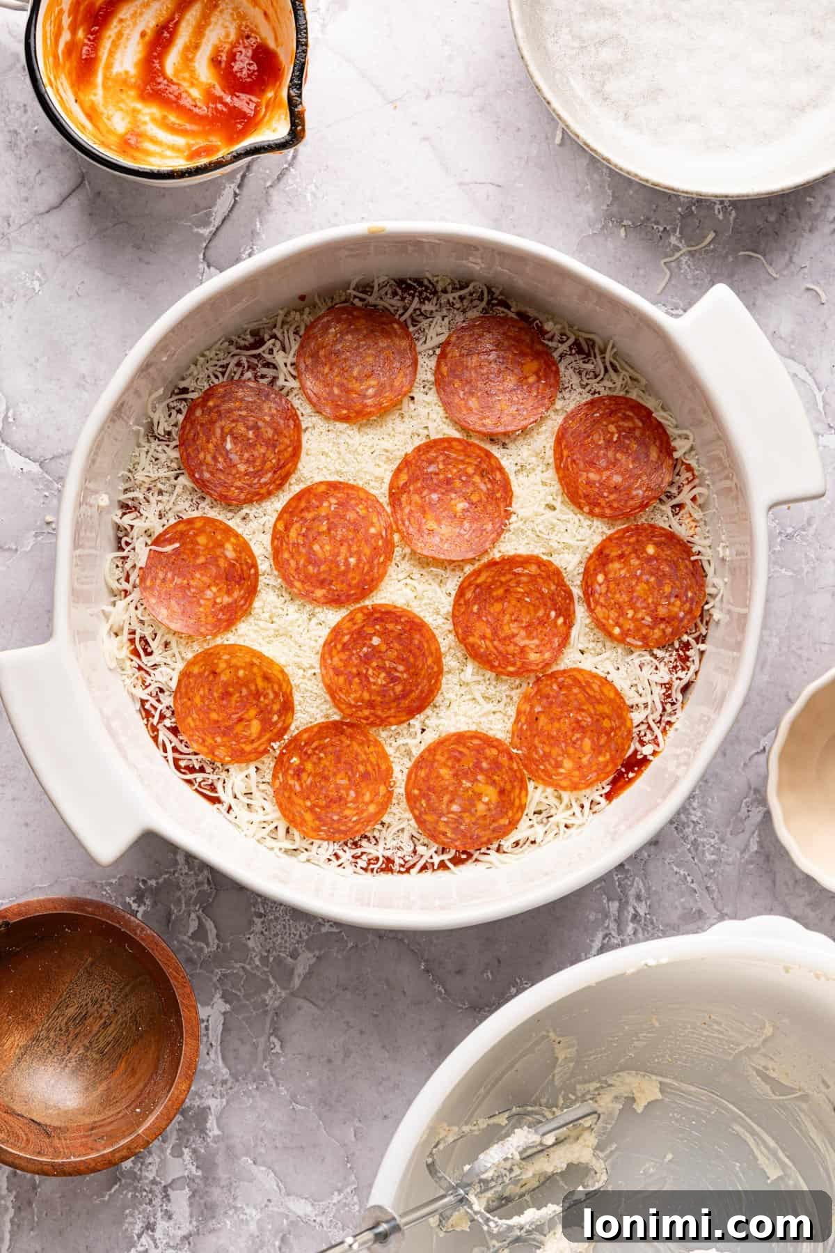 Unbaked high protein pizza dip in a baking dish, beautifully layered with the cheese base, pizza sauce, shredded cheese, and a single, neat layer of pepperoni slices on top.