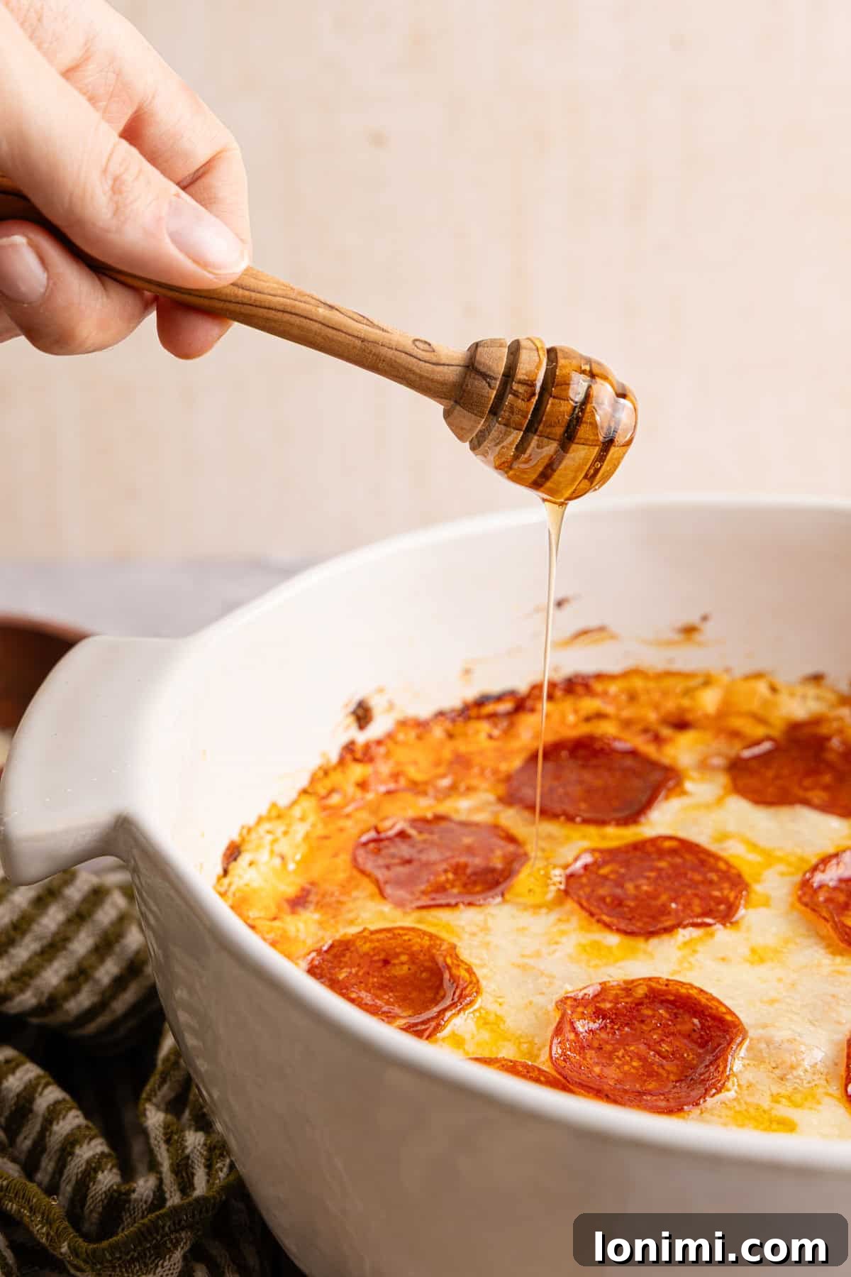 A hand carefully drizzling golden hot honey over the freshly baked high protein pizza dip, creating a tempting sweet and spicy glaze.