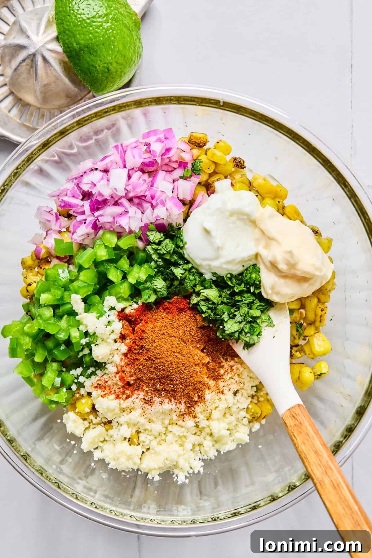 Street corn mixture being stirred with red onion, jalapeño, cilantro, mayo, Greek yogurt, lime juice, spices, and cotija cheese in a glass bowl.