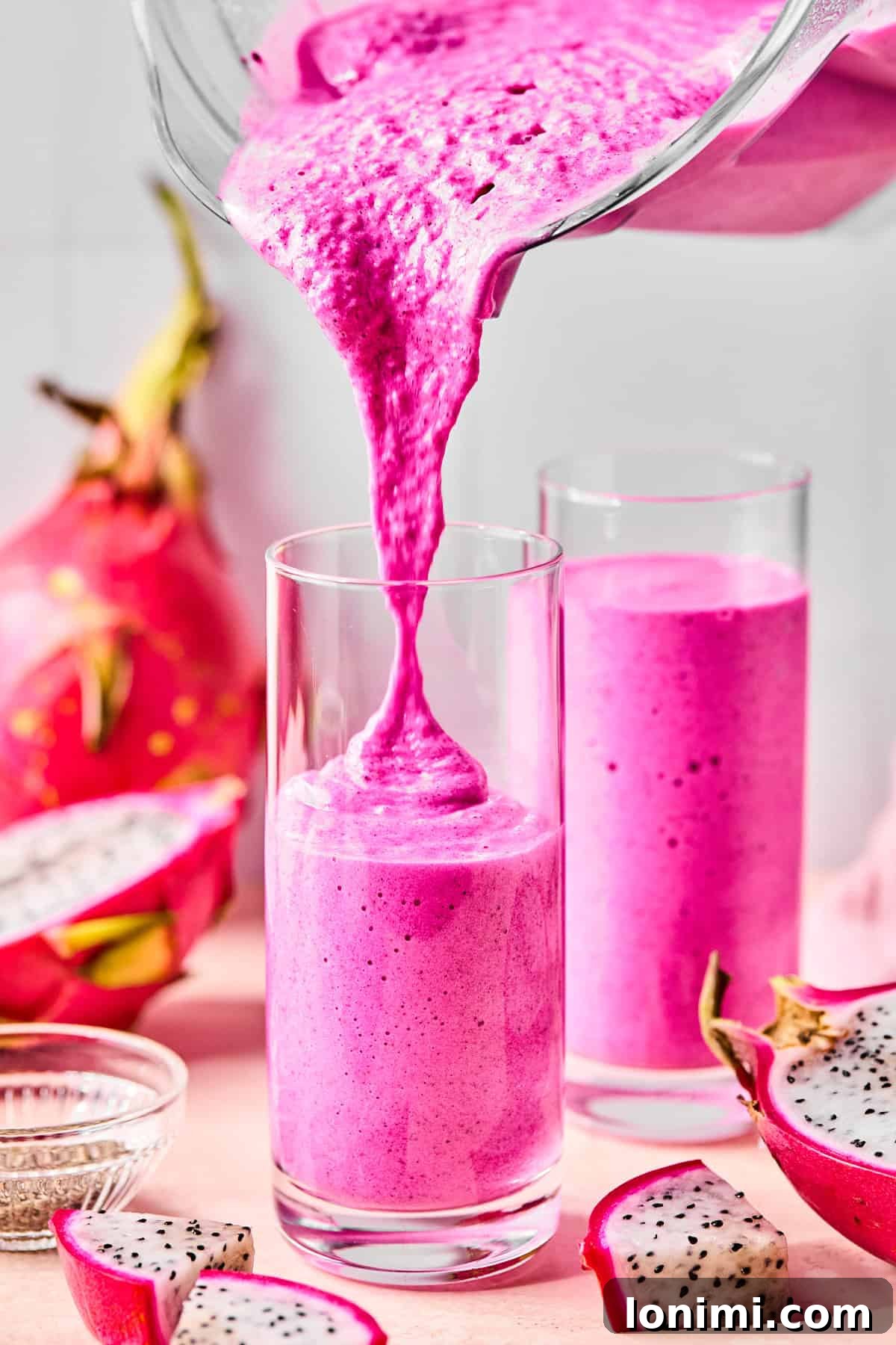 Dragonfruit smoothie being poured into a tall glass with another full glass behind it and sliced dragonfruit scattered around.