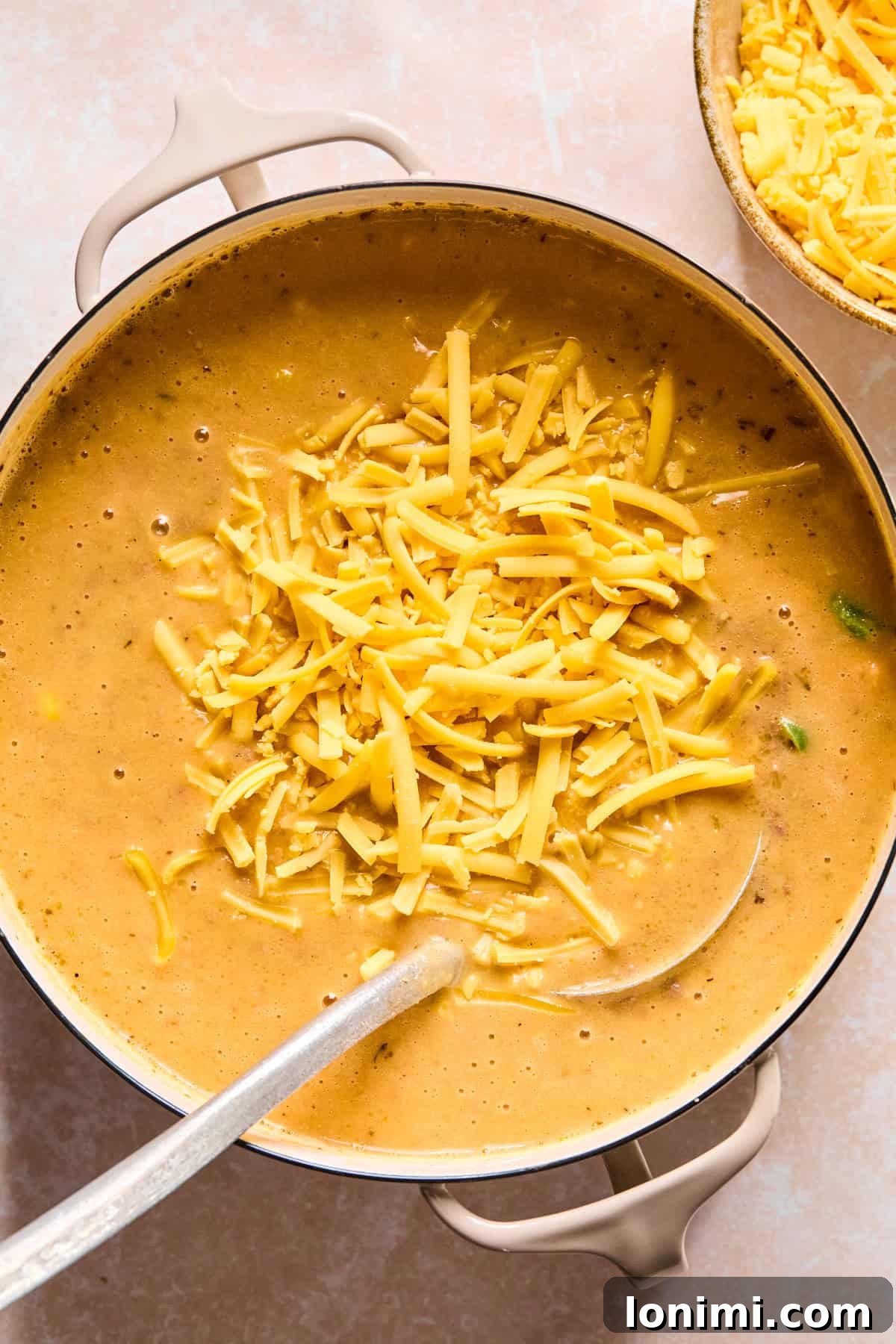 Shredded cheddar cheese being stirred into the hot soup with a ladle until smooth and melted.