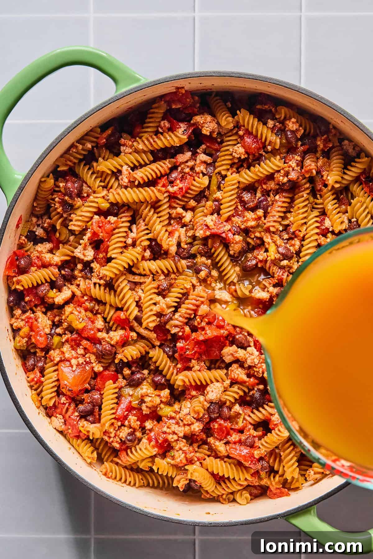 Easy Gluten-Free One Pot Taco Pasta 6 Chicken broth being poured into the pot with the taco seasoning, tomato paste, diced tomatoes, green chiles, black beans, and pasta.