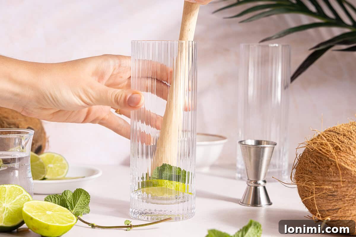 A lime wedge being gently muddled with fresh mint leaves in a tall cocktail glass