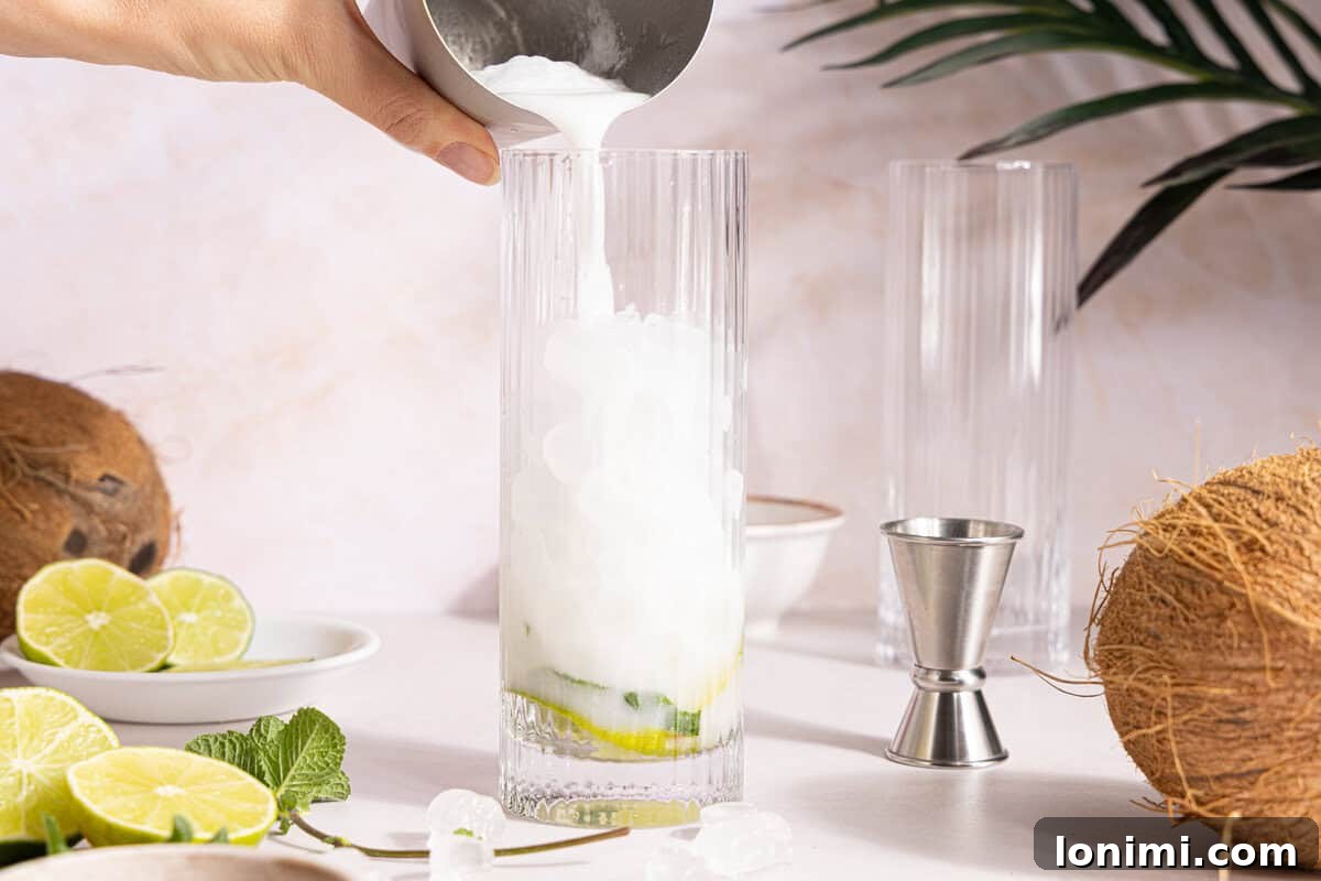 The creamy mixture of rum and cream of coconut being poured over ice in a cocktail glass with muddled mint and lime