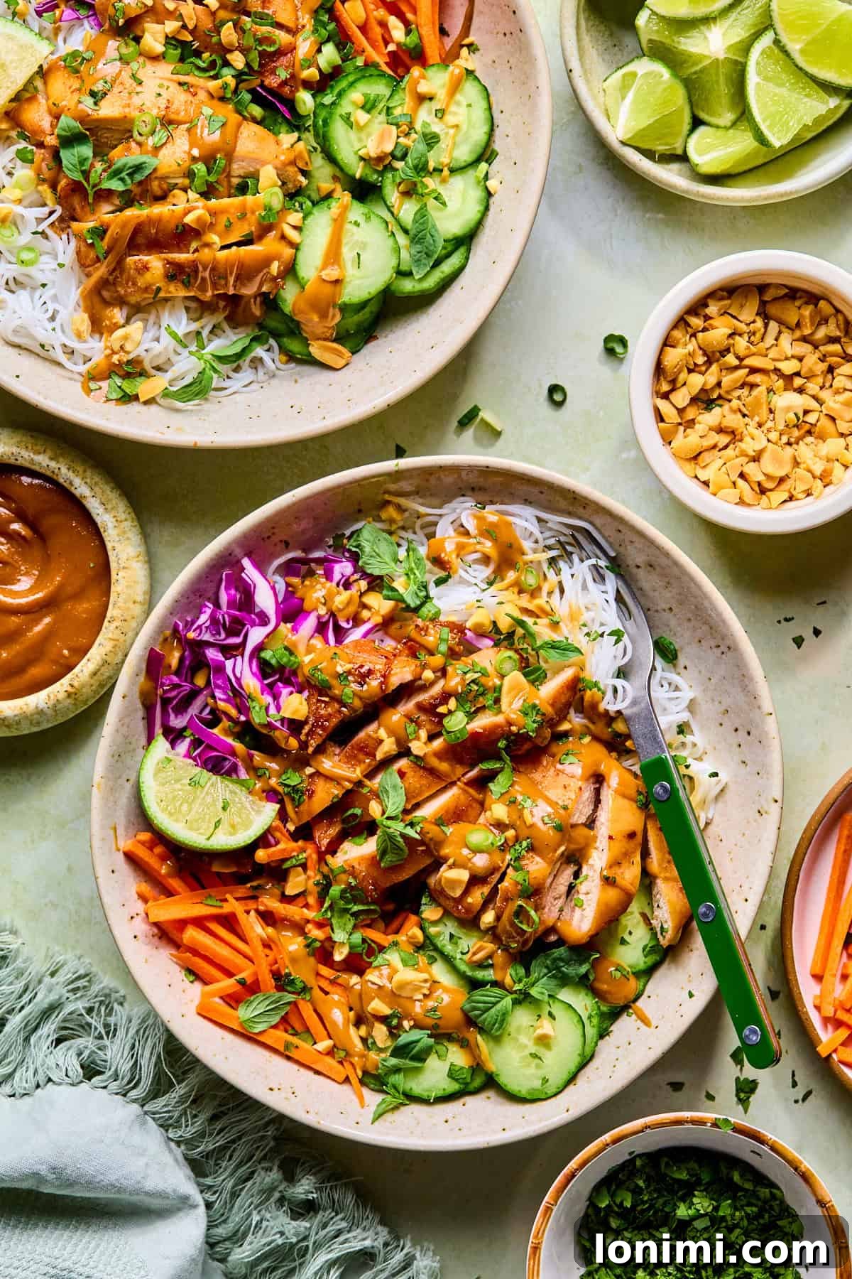 Two spring roll bowls topped with peanut sauce, surrounded by peanuts, fresh limes, cilantro, and extra peanut sauce.