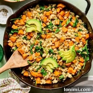 an egg-free breakfast hash with sweet potatoes, ground turkey, and spinach in a cast iron skillet topped with avocado with a wooden spoon