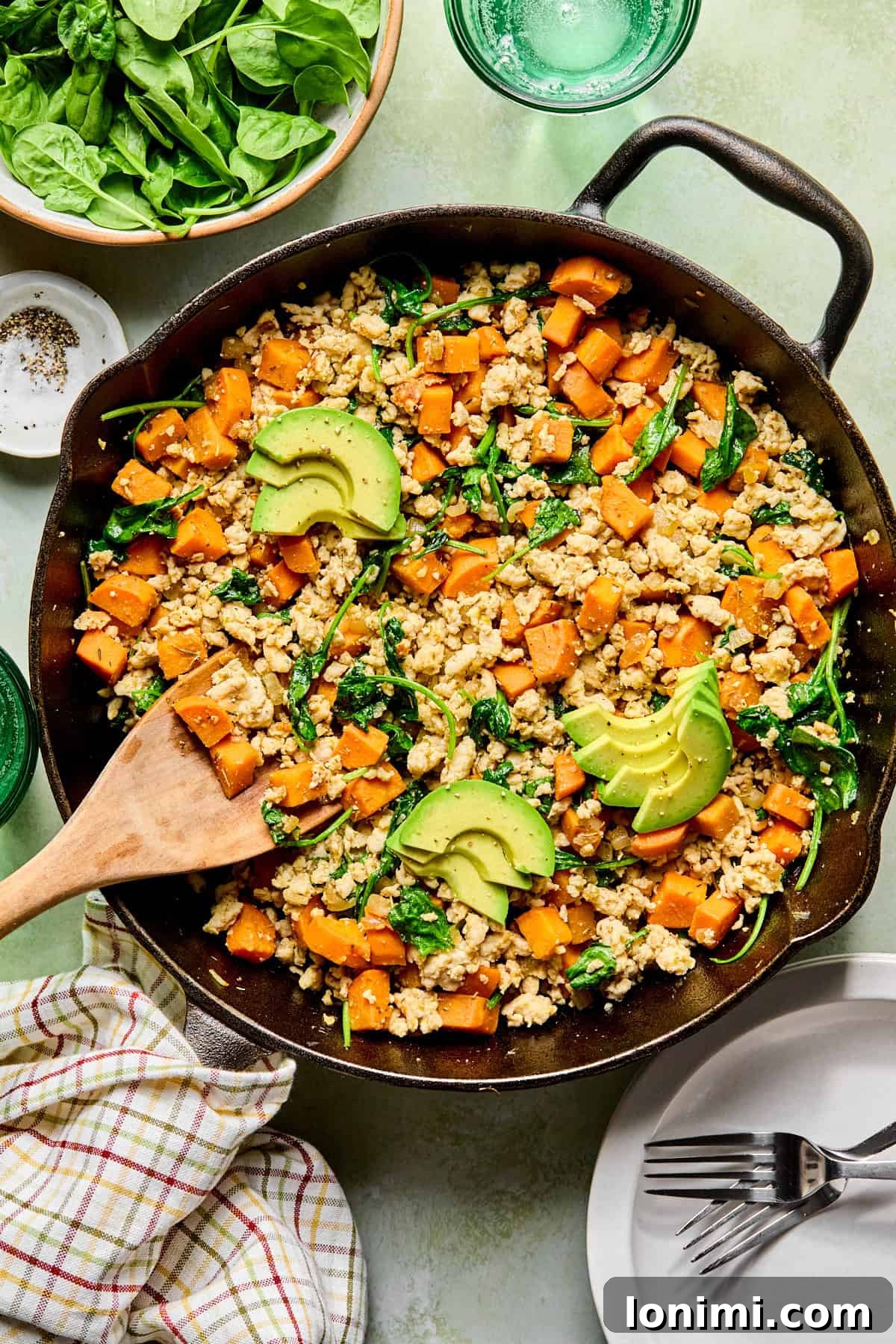 an egg-free breakfast hash with sweet potatoes, ground turkey, and spinach in a cast iron skillet topped with avocado with a wooden spoon